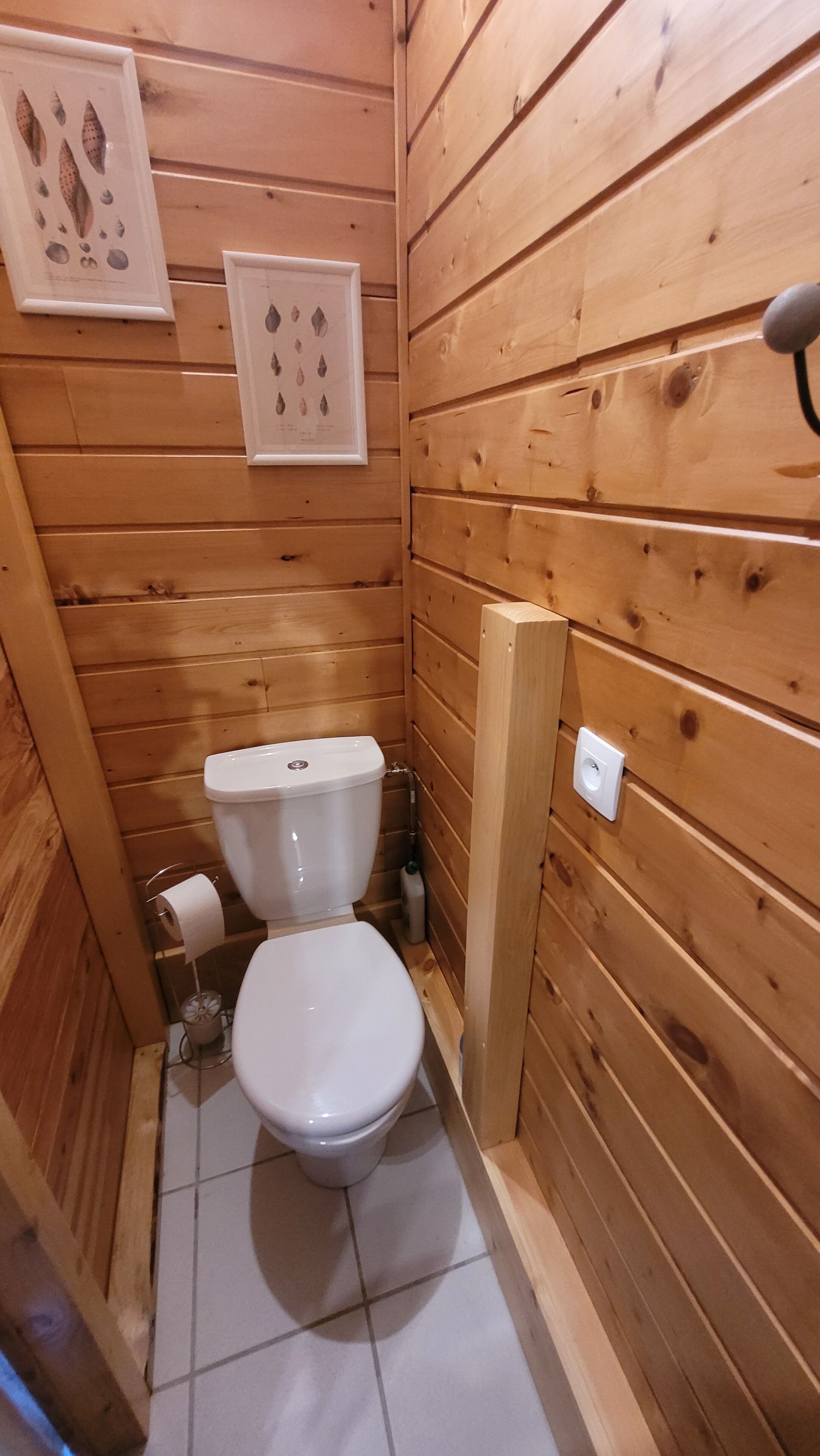 Ein kleines Badezimmer mit Holzvertäfelung, einer Toilette, einem Toilettenpapierhalter und einem an der Wand befestigten Lichtschalter.