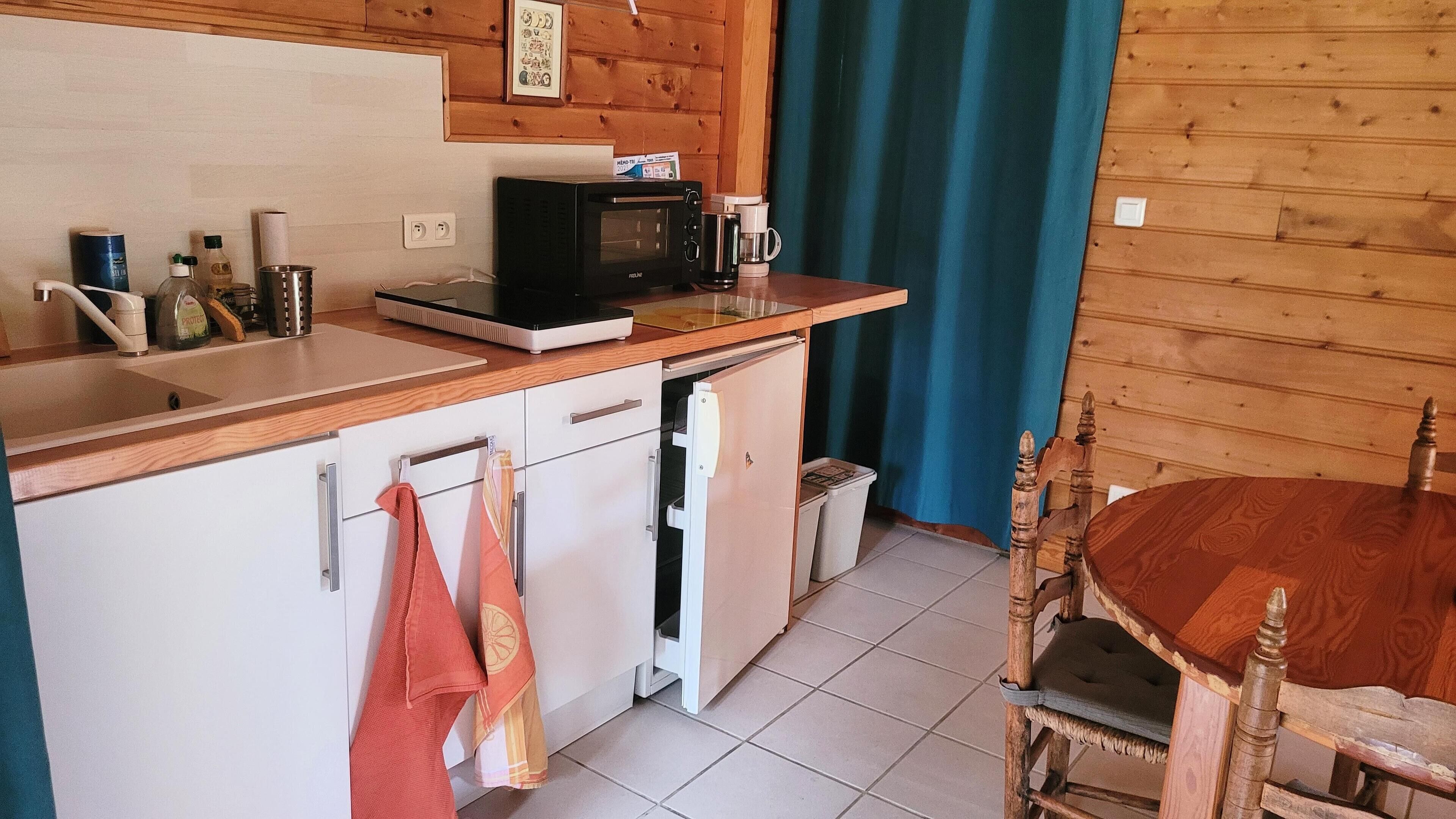 Une cuisine compacte équipée d’un four à micro-ondes, d’une machine à café et d’un petit réfrigérateur. On y trouve une table à manger en bois avec des chaises, et un rideau bleu en arrière-plan.