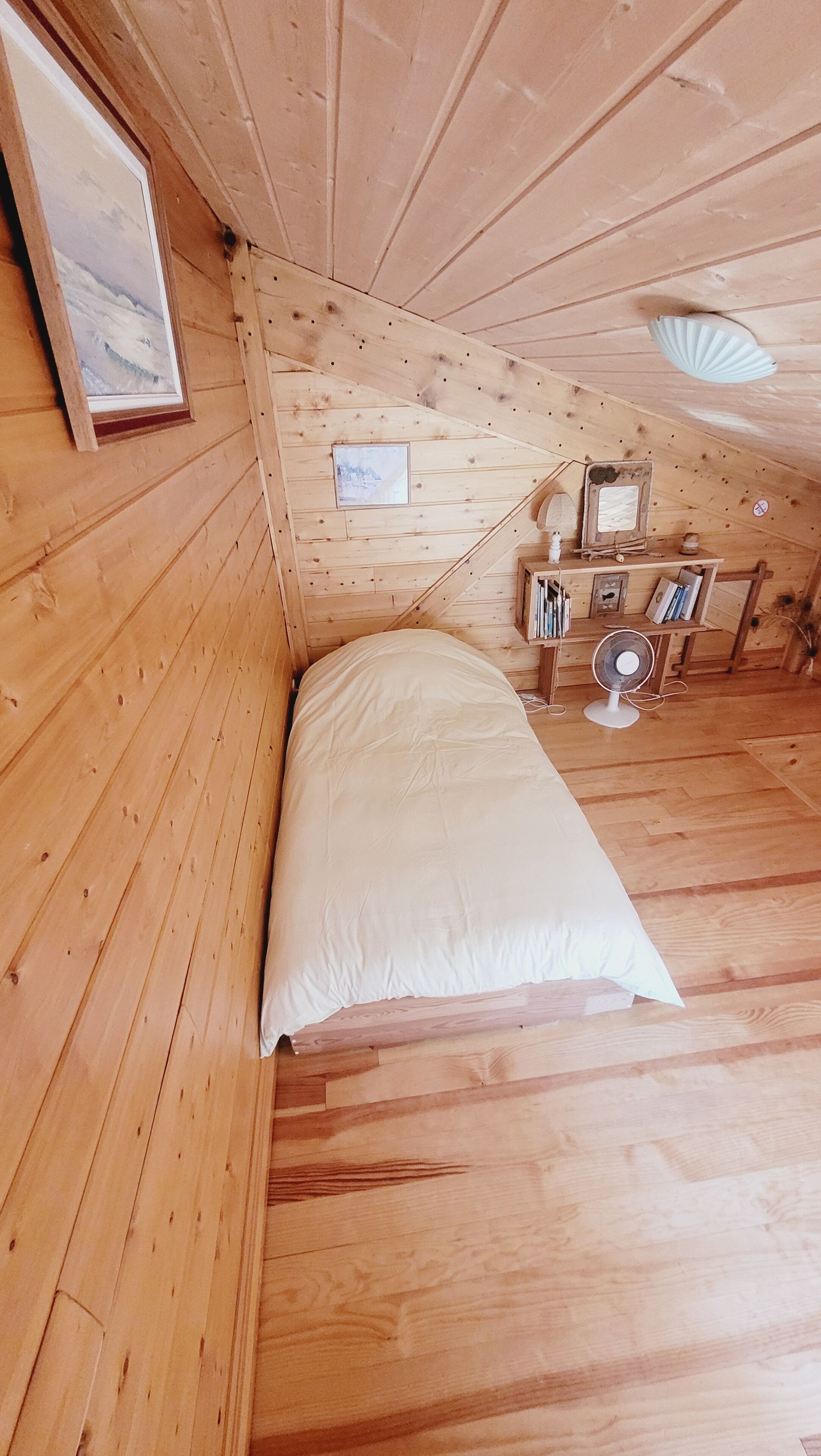 Una habitació de fusta amb un llit, una taula petita amb llibres, un ventilador i una estanteria amb diferents objectes.