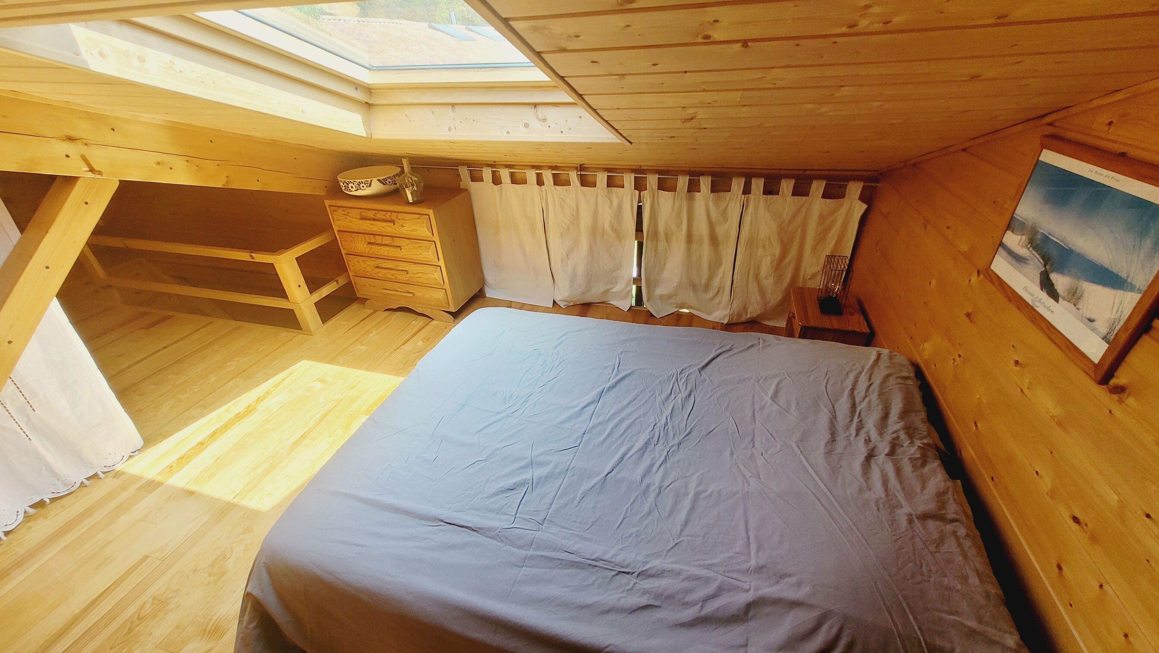 A cozy attic bedroom with a skylight, wooden walls, a bed, a nightstand, and a painting.