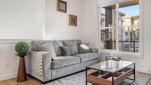 A modern living room with a grey sofa, a glass coffee table, and a large window offering a view of neighboring buildings. - King Bed » Garage » AC » Full Kitchen » 65` (Calgary)