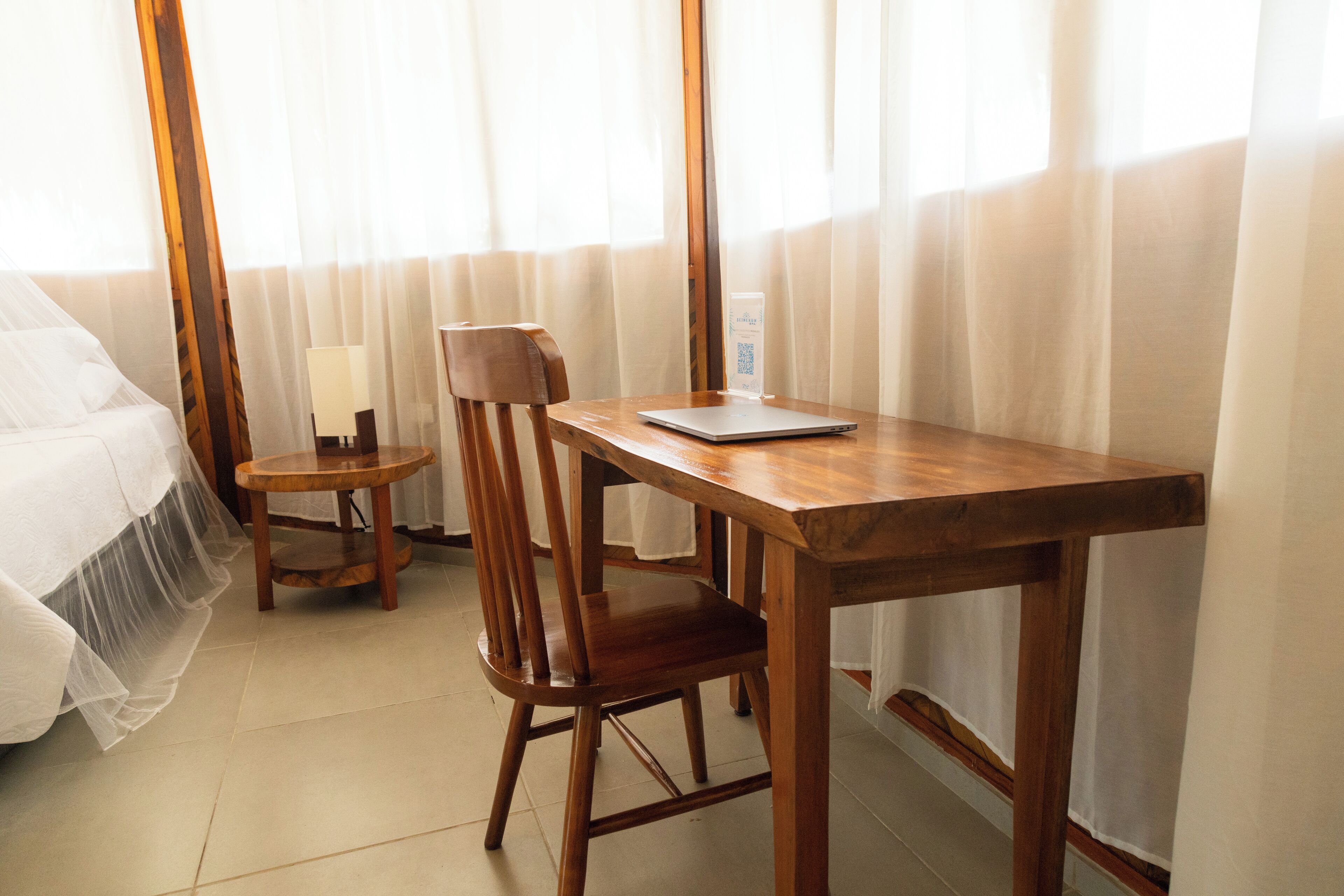 A hotel room with a wooden desk, a chair, a bed with white bedding, a small table with a lamp, and sheer curtains on the windows.