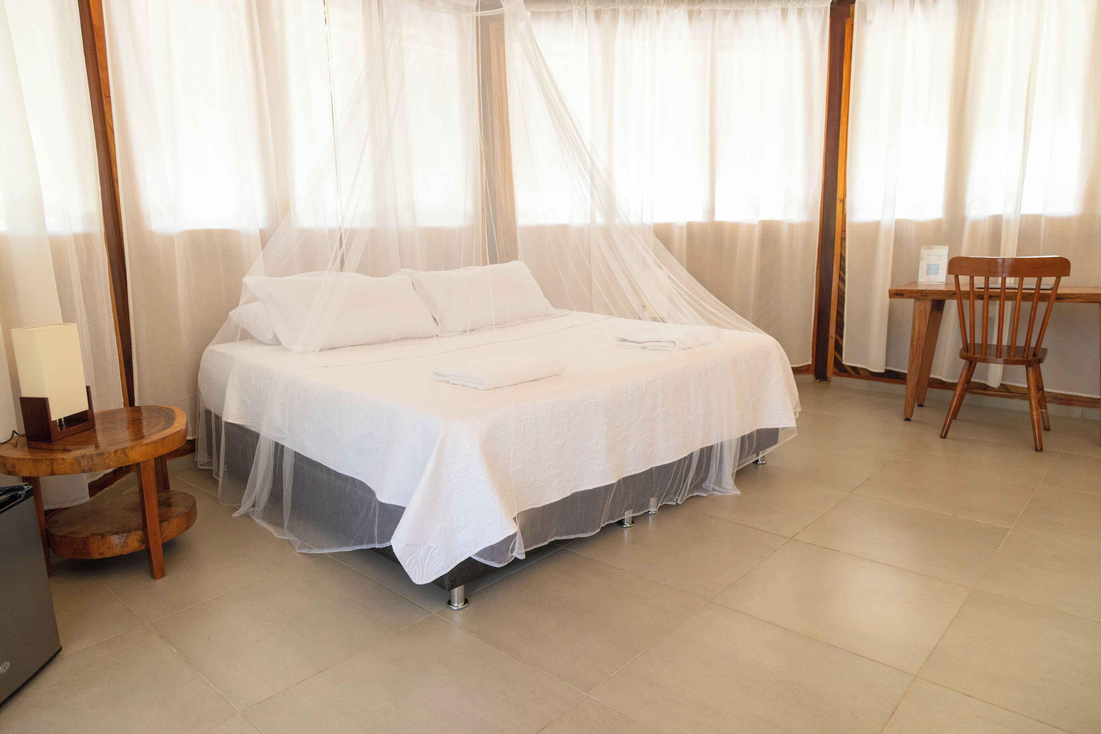 A neatly made bed with white linens, a wooden nightstand with a lamp, and a chair by the window.