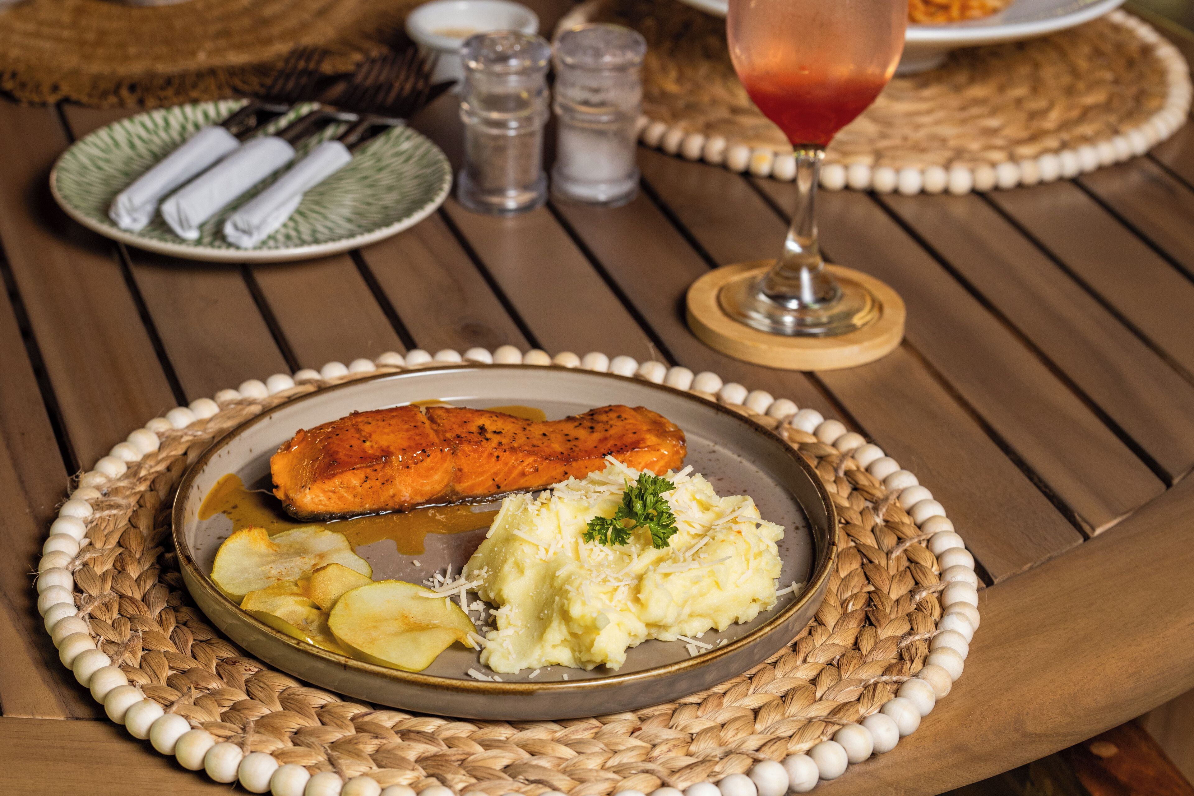 A wooden table with a woven placemat, a plate with salmon, mashed potatoes, and apple slices, and a glass of red wine.