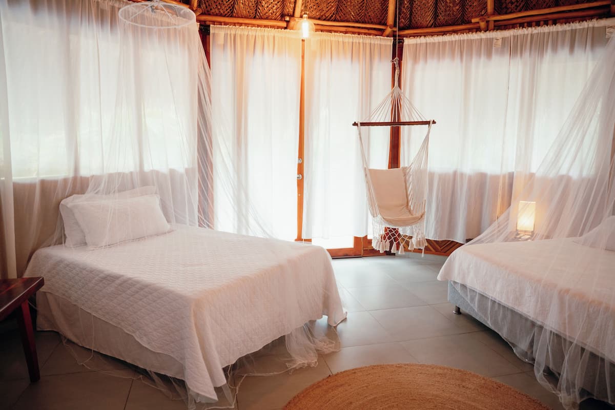 A room with two beds, a hanging chair, and a large window with sheer curtains.