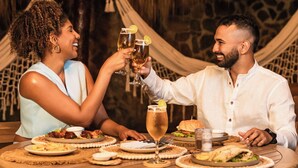 Un couple trinque avec des boissons autour d’une table ronde en bois, dans un cadre rustique agrémenté d’un toit de chaume et d’un hamac en arrière-plan.
