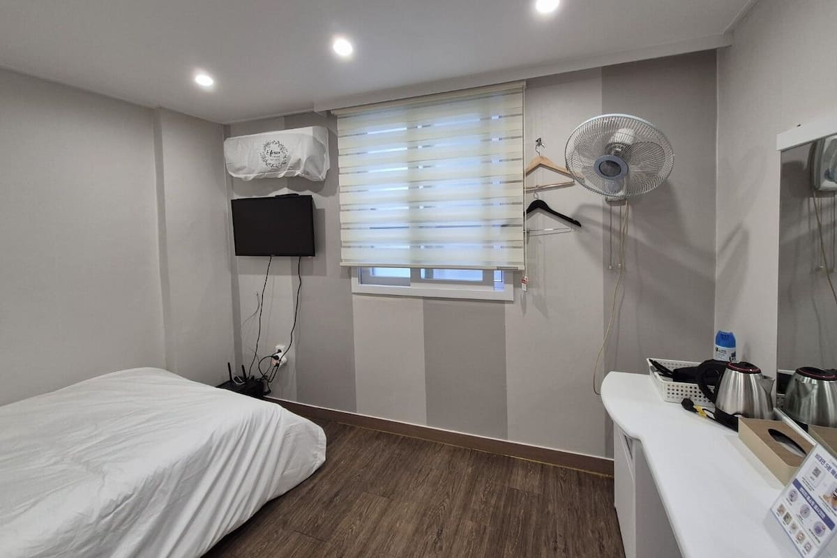 A hotel room with a bed, a TV mounted on the wall, a window with blinds, a standing fan, and a small table with a kettle and a basket.