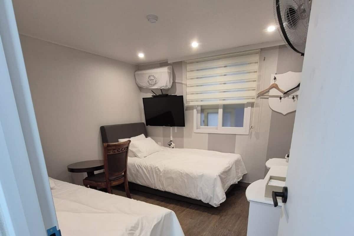 A hotel room with two beds, a TV, a fan, a window with blinds, and a small table.