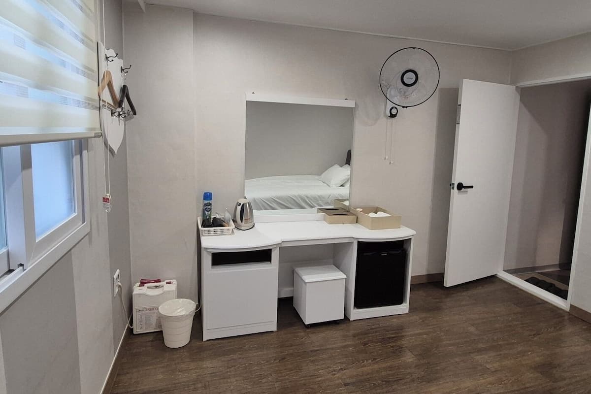 A compact bedroom with a bed, a desk, a small cabinet, a fan, and a door.