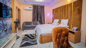 A modern bedroom with a large bed, a grey sofa, a wooden nightstand, and a TV mounted on the wall. - Southern Altantic  Hotels and Apartments (Yenagoa)