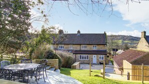 A stone house with a tiled roof, a patio with metal chairs, and a fenced garden.