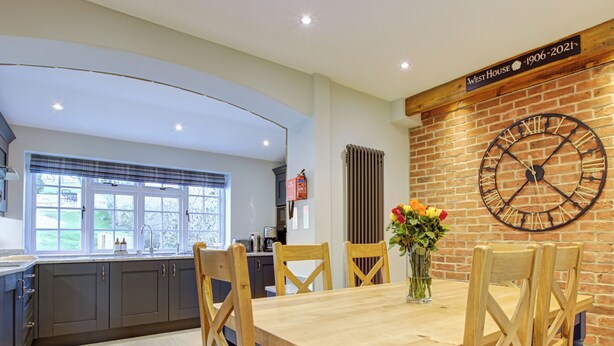 A modern kitchen with a large dining table, wooden chairs, and a clock on the wall.