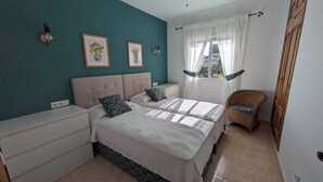 A bedroom with a teal wall, two beds with white bedding, a nightstand, a wicker chair, and a window with sheer curtains. - Villa Carolina. Costa Nova, Jávea (Xàbia)