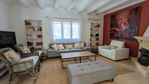 A living room with a sofa, ottoman, and chairs, a television, and bookshelves. - Villa Carolina. Costa Nova, Jávea (Xàbia)