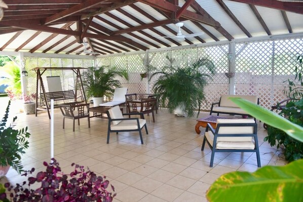 Ein überdachter Patio-Bereich mit Holzmöbeln, darunter Stühle und ein Tisch, umgeben von Pflanzen in Töpfen.