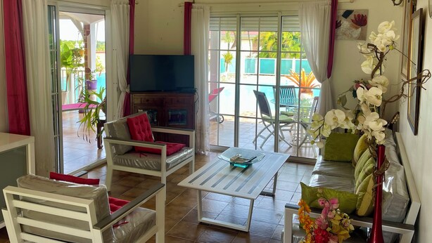 A living room with a TV, seating area, and a view of the pool. - Villa Horizon Caraïbes (Petit-Bourg)