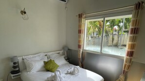 A neatly made bed with white linens and a green pillow, a bedside lamp, and a window with curtains. - Villa Horizon Caraïbes (Petit-Bourg)