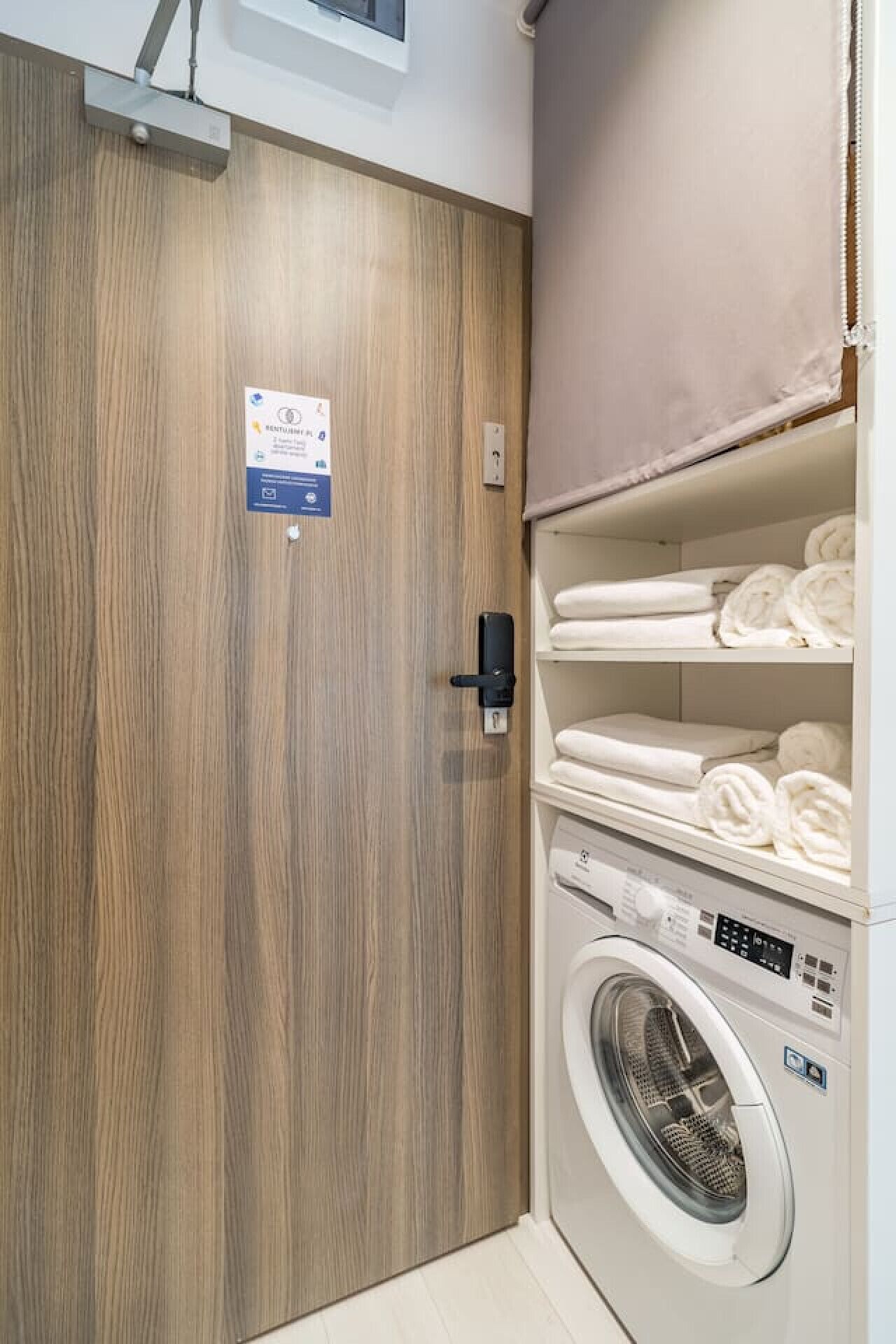 A laundry room with a wooden door, a wall-mounted washing machine, and shelves with folded towels.