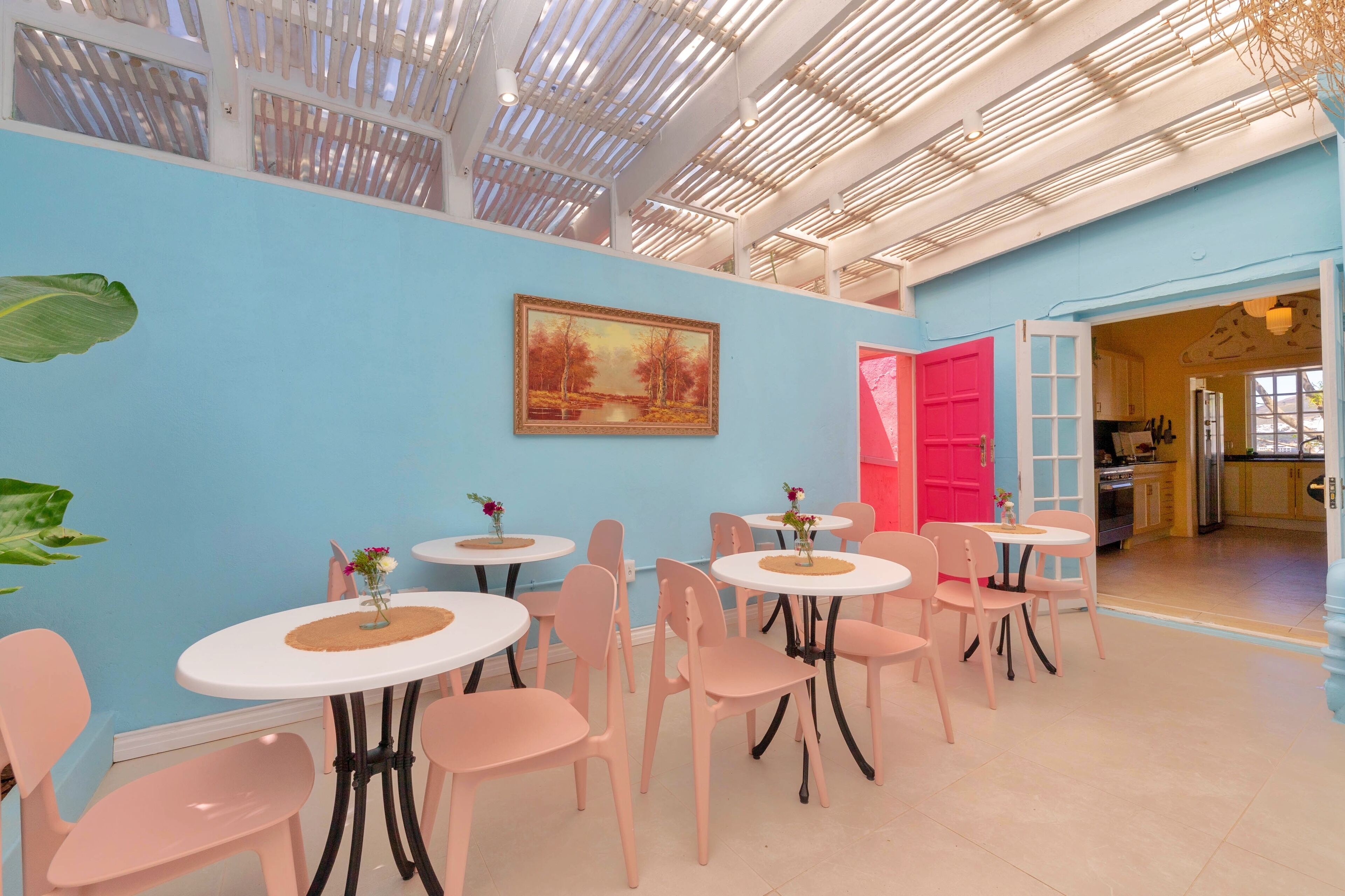 Une salle à manger lumineuse, avec des tables rondes et des chaises roses, un grand tableau au mur et une cuisine visible à travers une porte ouverte.
