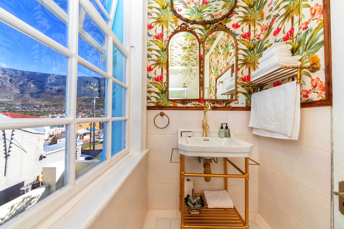 Un bagno con un'ampia finestra, un lavabo con dettagli dorati e uno specchio con carta da parati floreale.