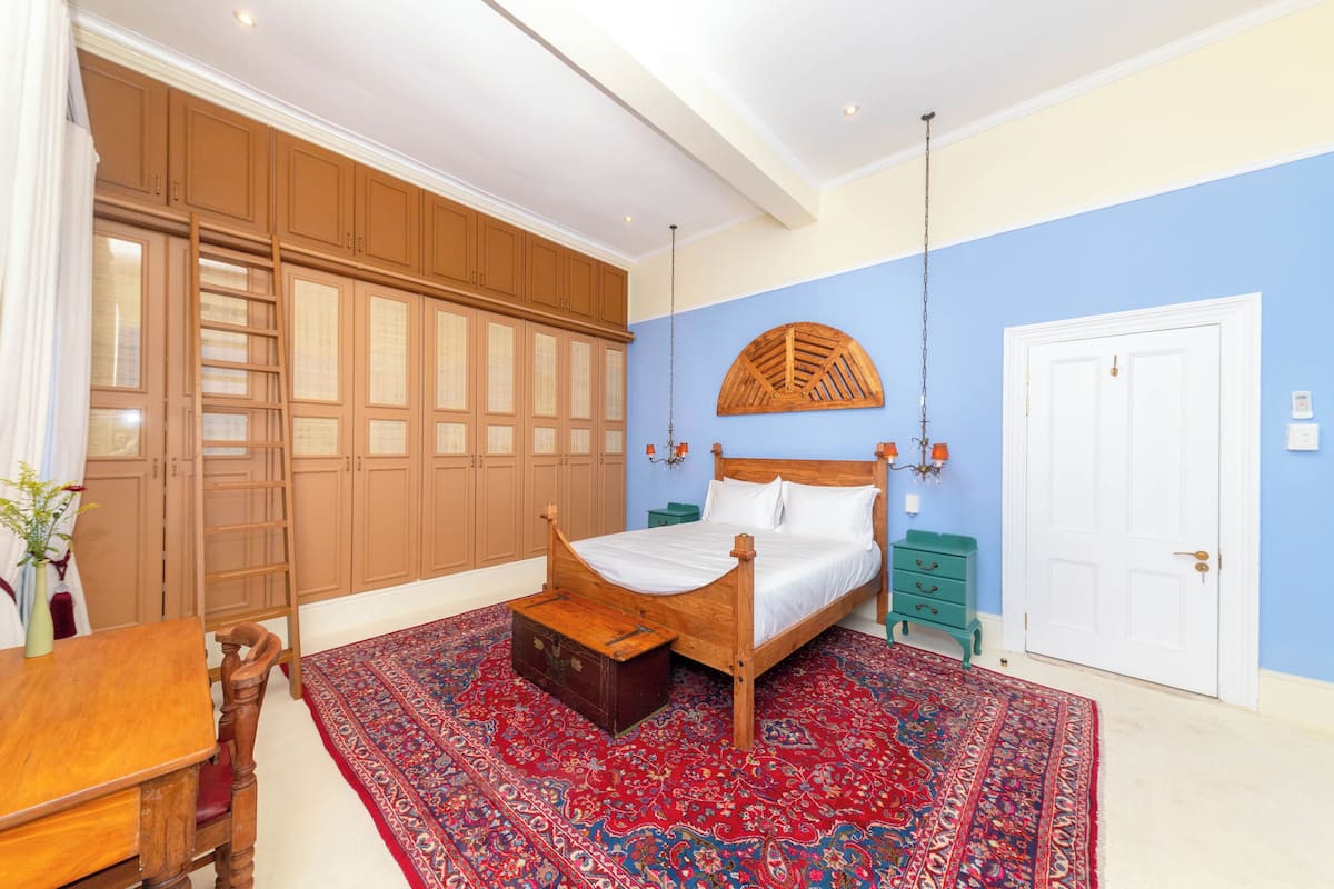 Une chambre à coucher avec un lit en bois, un tapis rouge à motifs, une armoire en bois et une porte blanche.