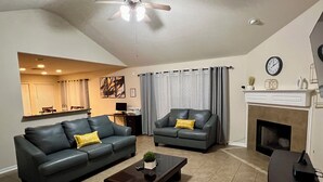 A living room with two dark sofas, a coffee table, a fireplace, a ceiling fan, and a kitchen area in the background. - Cozy Home in Spring,TX with AC /WiFi / Laundry . 20 mins from IAH Airport (Spring)