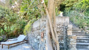 A stone staircase leading up to a stone wall with a tree growing through it. There are two lounge chairs with white cushions on a tiled patio area. - La Spiaggia (Moneglia)