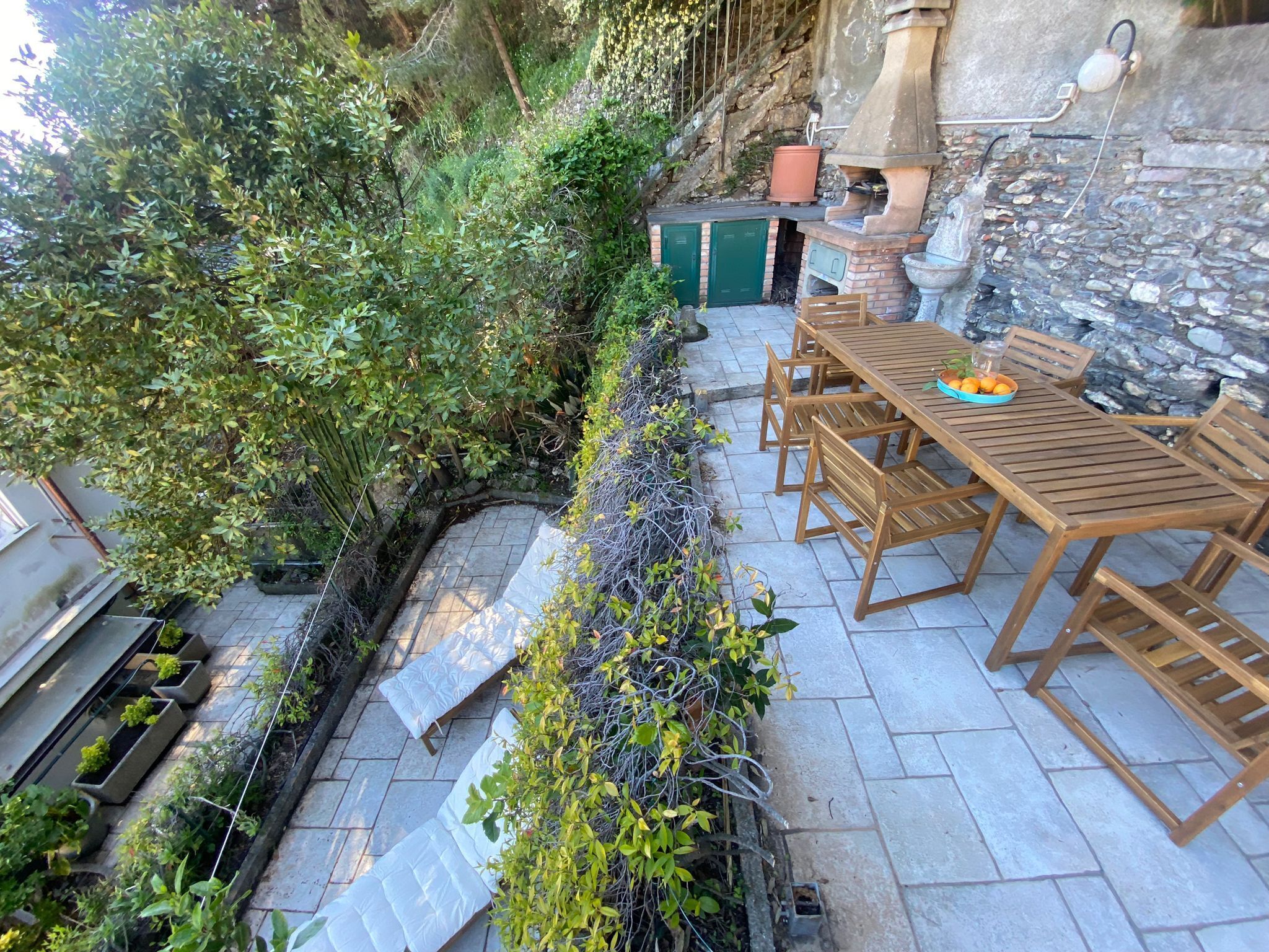 Eine Terrasse mit einem Steintisch und Holzstühlen, umgeben von Grünpflanzen und einer Steinmauer.
