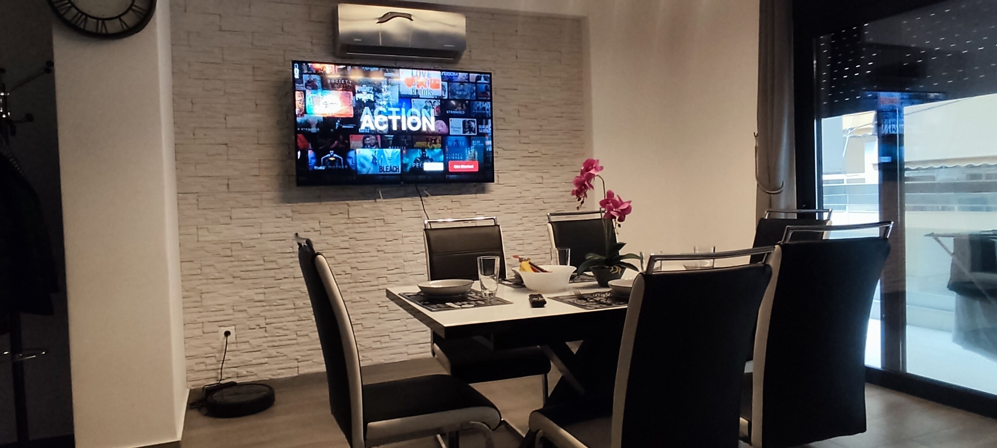 Une chambre moderne avec un mur en briques, un téléviseur à écran plat diffusant différentes chaînes, une table à manger avec des chaises et une orchidée en pot.