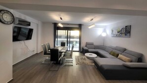 Un salon moderne comprenant un coin repas, un canapé gris, un téléviseur à écran plat et une grande fenêtre donnant sur des immeubles.
