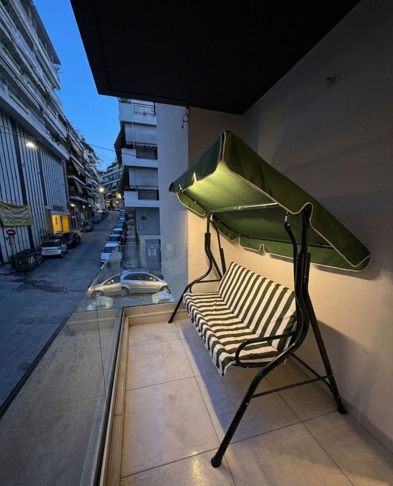 Un balcon avec un canapé à rayures et un auvent vert, donnant sur une rue bordée de voitures garées et d’immeubles.
