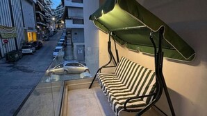Un balcon avec un canapé à rayures et un auvent vert, donnant sur une rue bordée de voitures garées et d’immeubles.