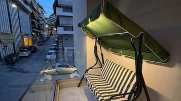 Un balcon avec un canapé à rayures et un auvent vert, donnant sur une rue bordée de voitures garées et d’immeubles.