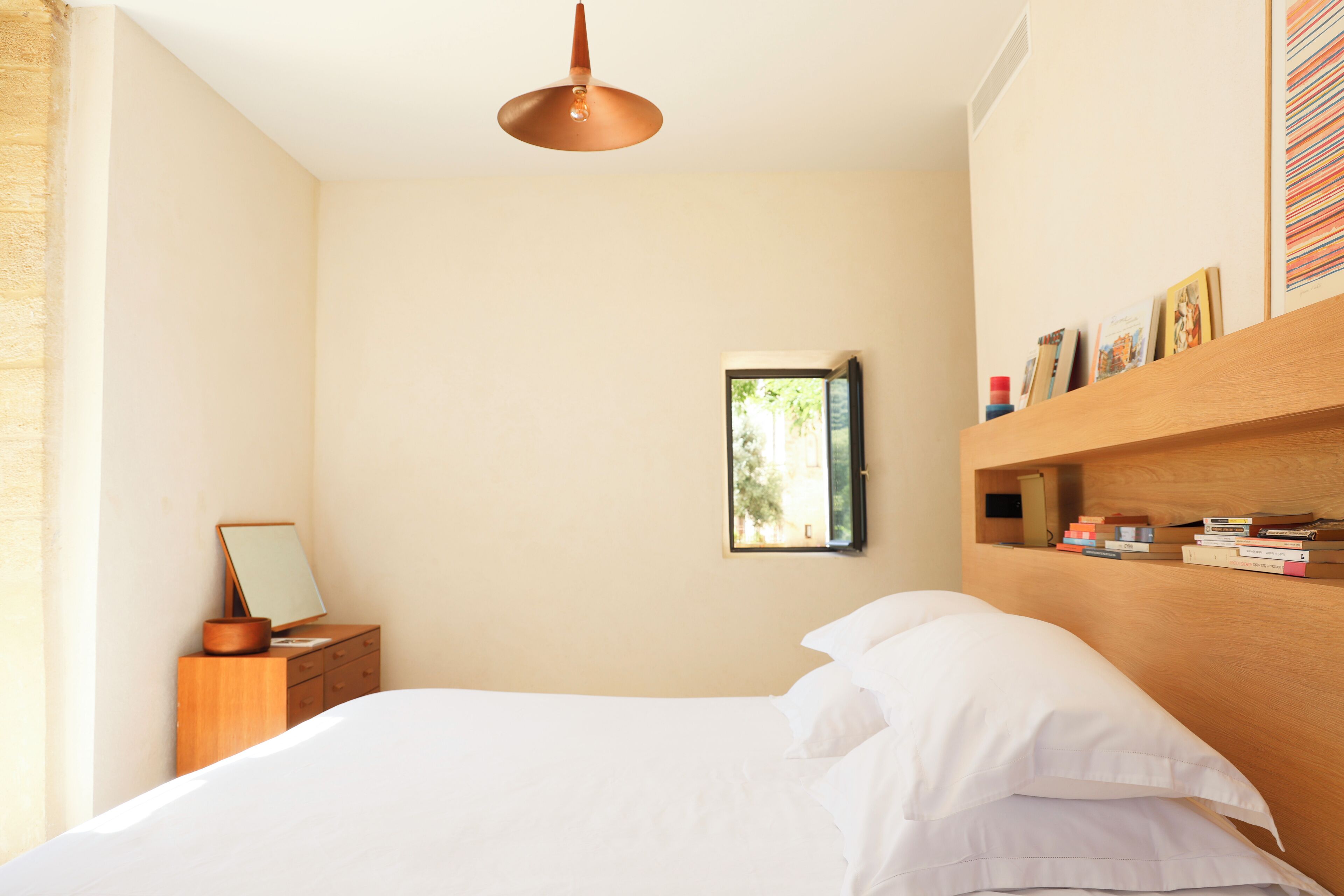Een slaapkamer met een bed, een houten nachtkastje, een spiegel, een plank met boeken en een raam.