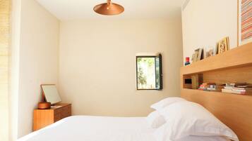 Een slaapkamer met een bed, een houten nachtkastje, een spiegel, een plank met boeken en een raam.