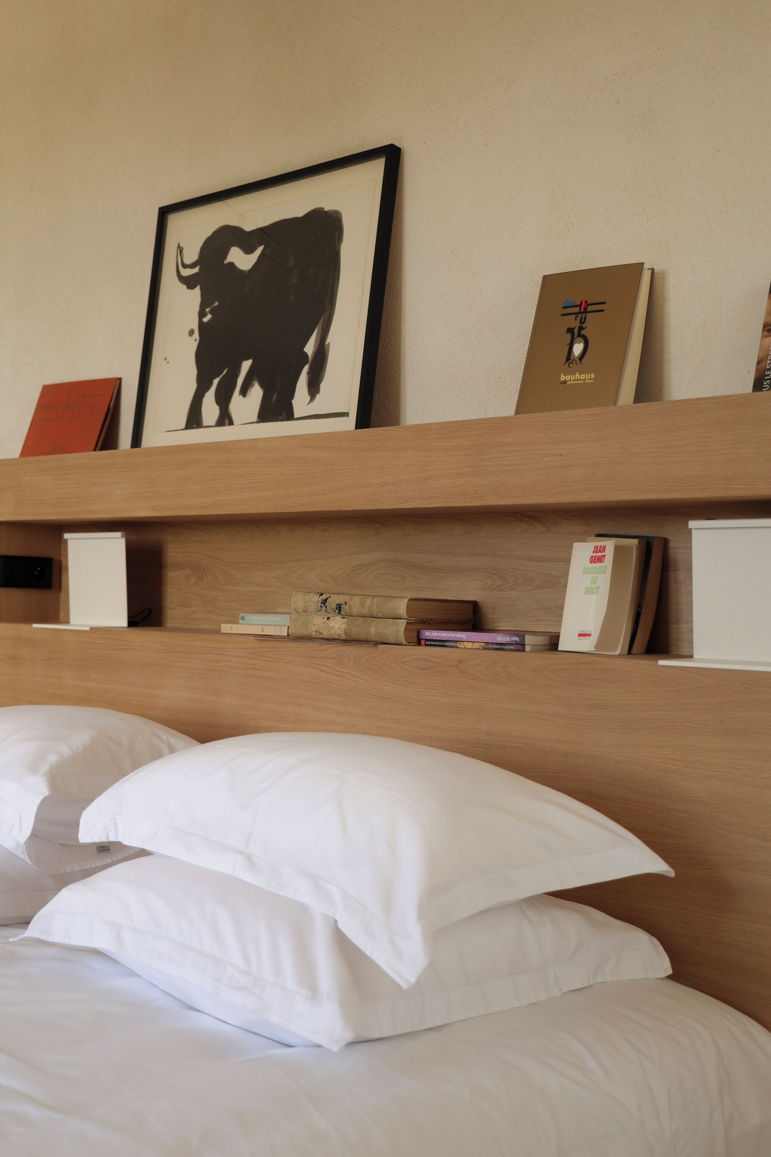 Una habitación de hotel con una cama, almohadas, un cabecero de madera, una obra de arte enmarcada de un toro y una estantería con libros y una pequeña caja.