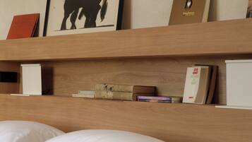 Una habitación de hotel con una cama, almohadas, un cabecero de madera, una obra de arte enmarcada de un toro y una estantería con libros y una pequeña caja.