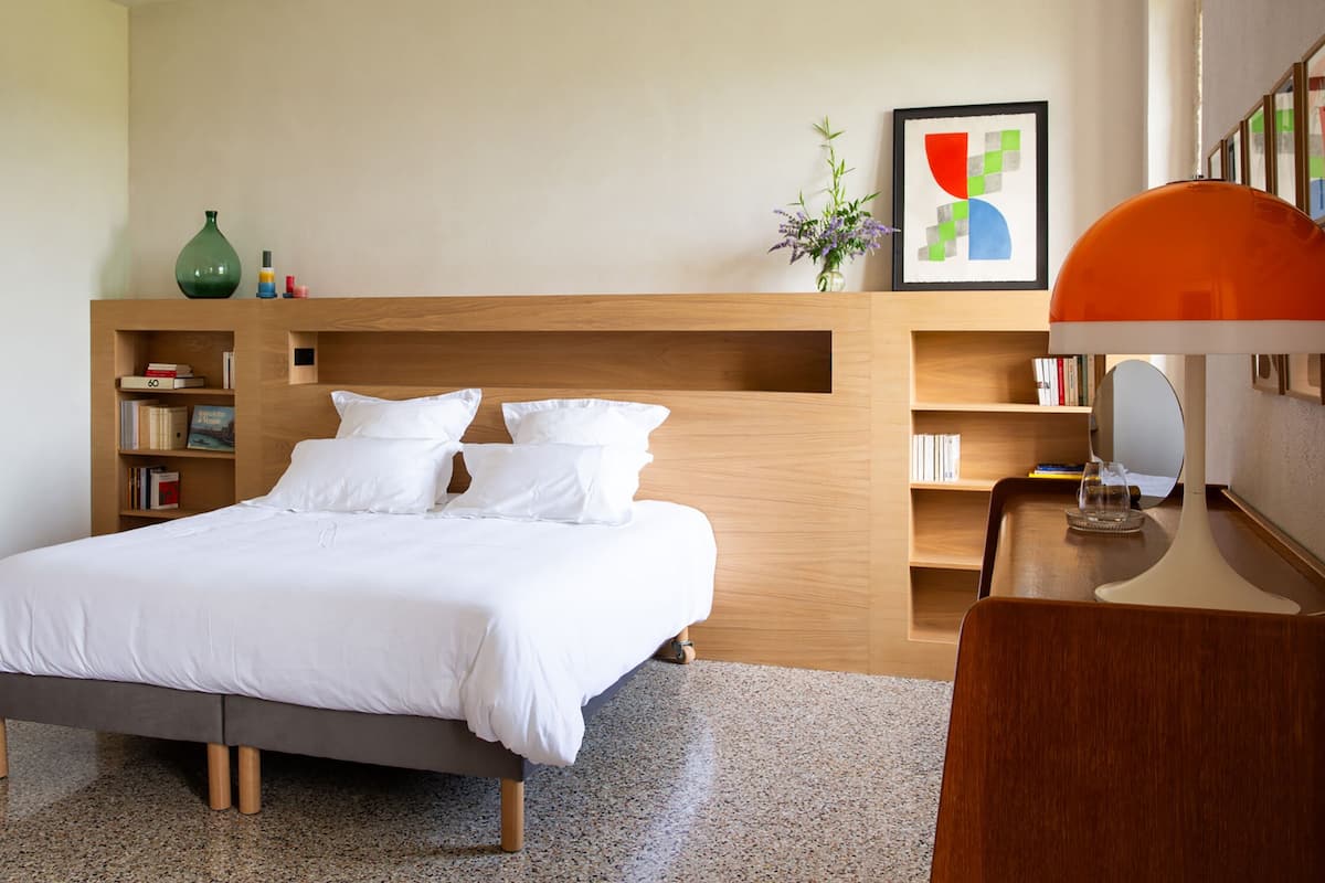 A modern bedroom with a bed, wooden headboard, bookshelves, a bedside table with a lamp, and a framed abstract painting.