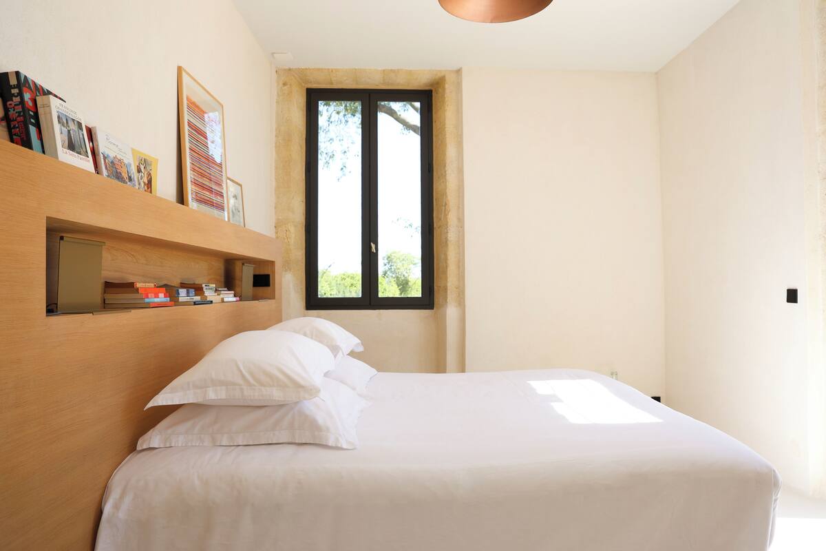 A bedroom with a bed, a wooden shelf with books, and a window with a view of greenery.