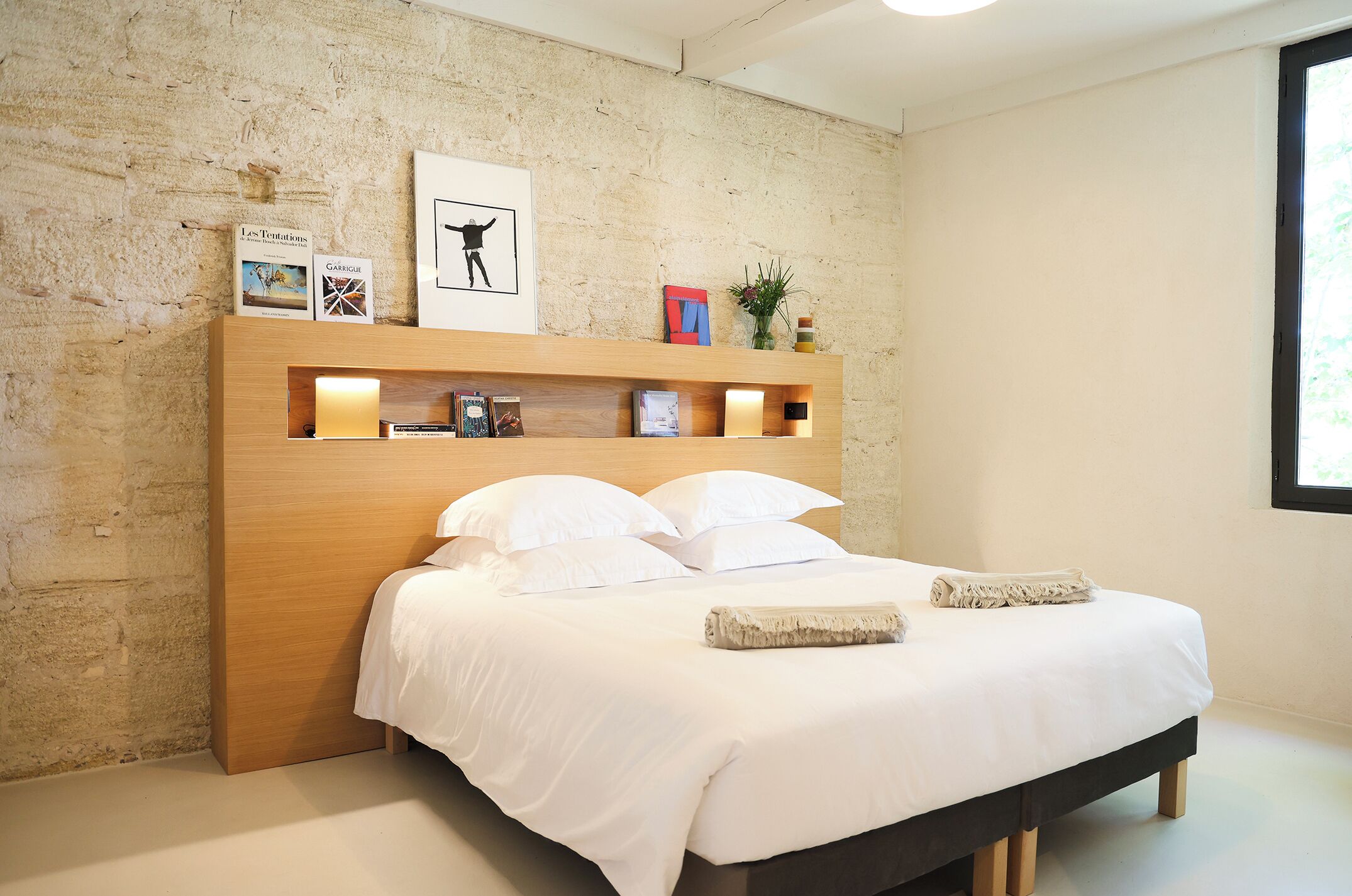Un dormitorio moderno con cabecera de madera, ropa de cama blanca y una estantería con libros y objetos decorativos.