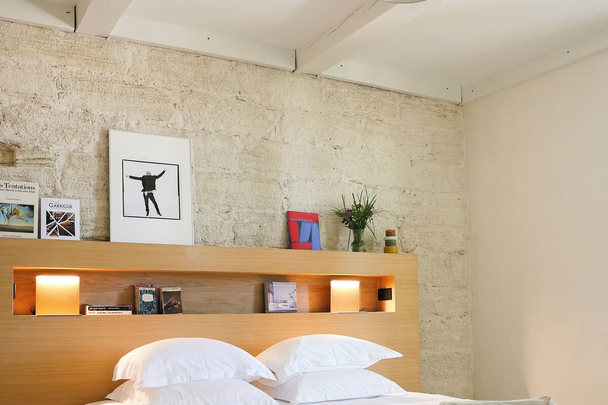 A modern bedroom with a wooden headboard, a bed with white linens, a pendant light, and a framed picture on the wall.