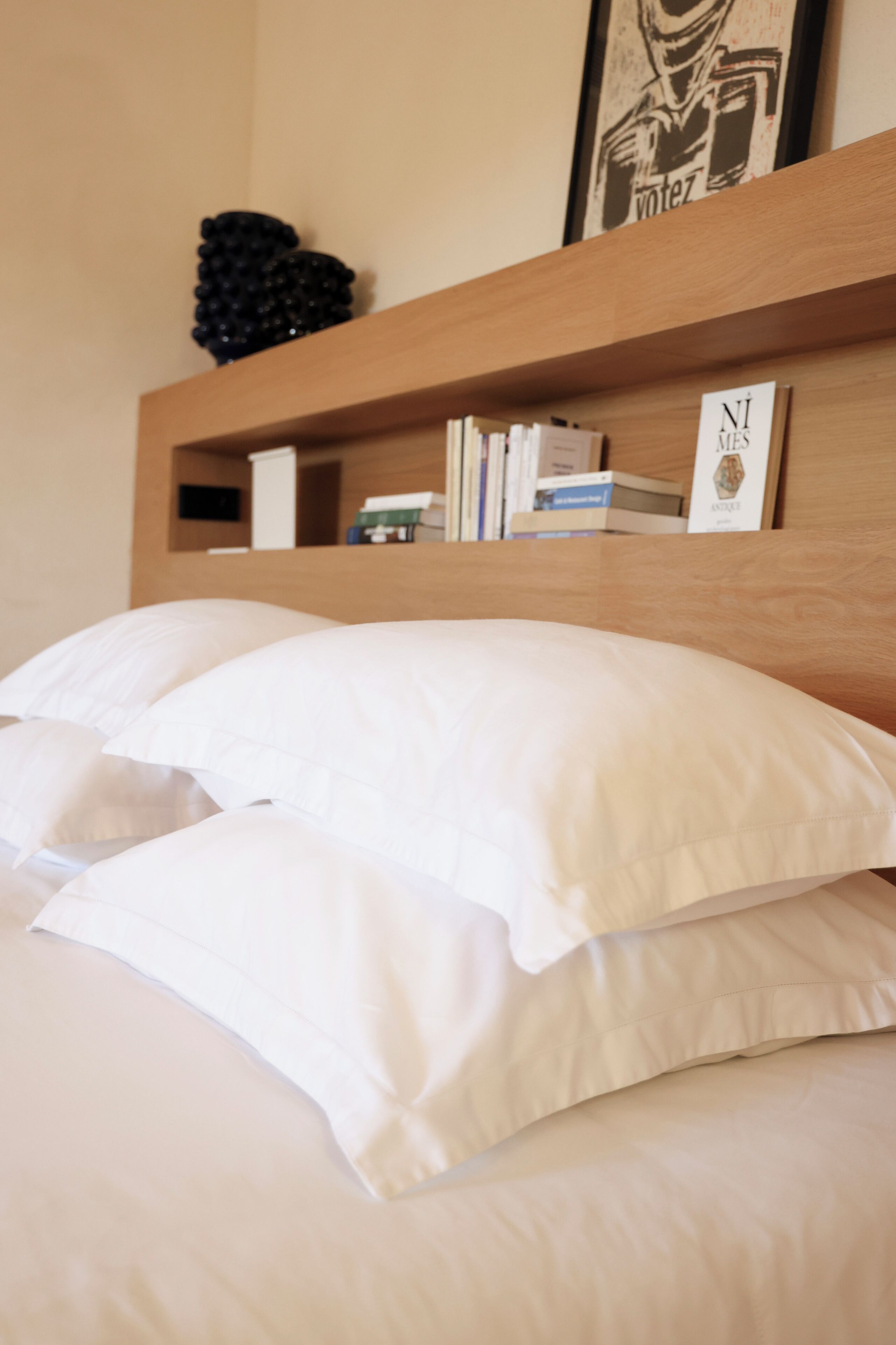 Una cama bien hecha con sábanas y almohadas blancas, un cabecero de madera y un estante superior con libros y un cuadro enmarcado.