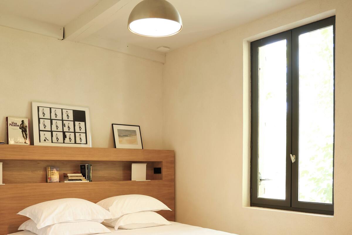 A bedroom with a bed, a window, a shelf with books and pictures, and a pendant light.