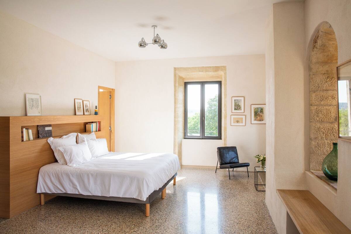 A bedroom with a bed, a wooden headboard, a bookshelf, a window, a chair, and a vase with flowers.
