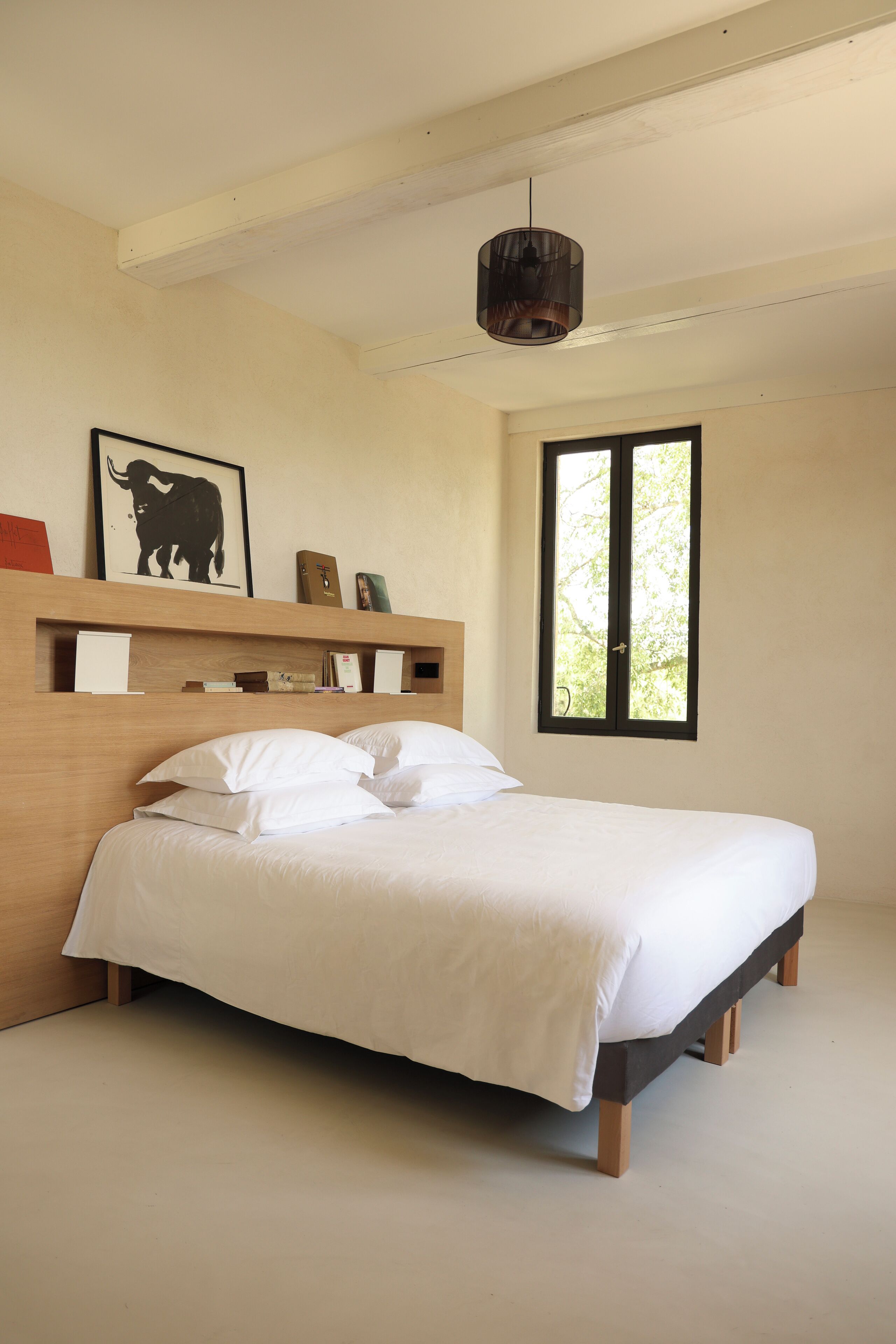 Une chambre à coucher avec un cadre de lit en bois, du linge de lit blanc, une œuvre d’art encadrée représentant un taureau et une fenêtre donnant sur des arbres.