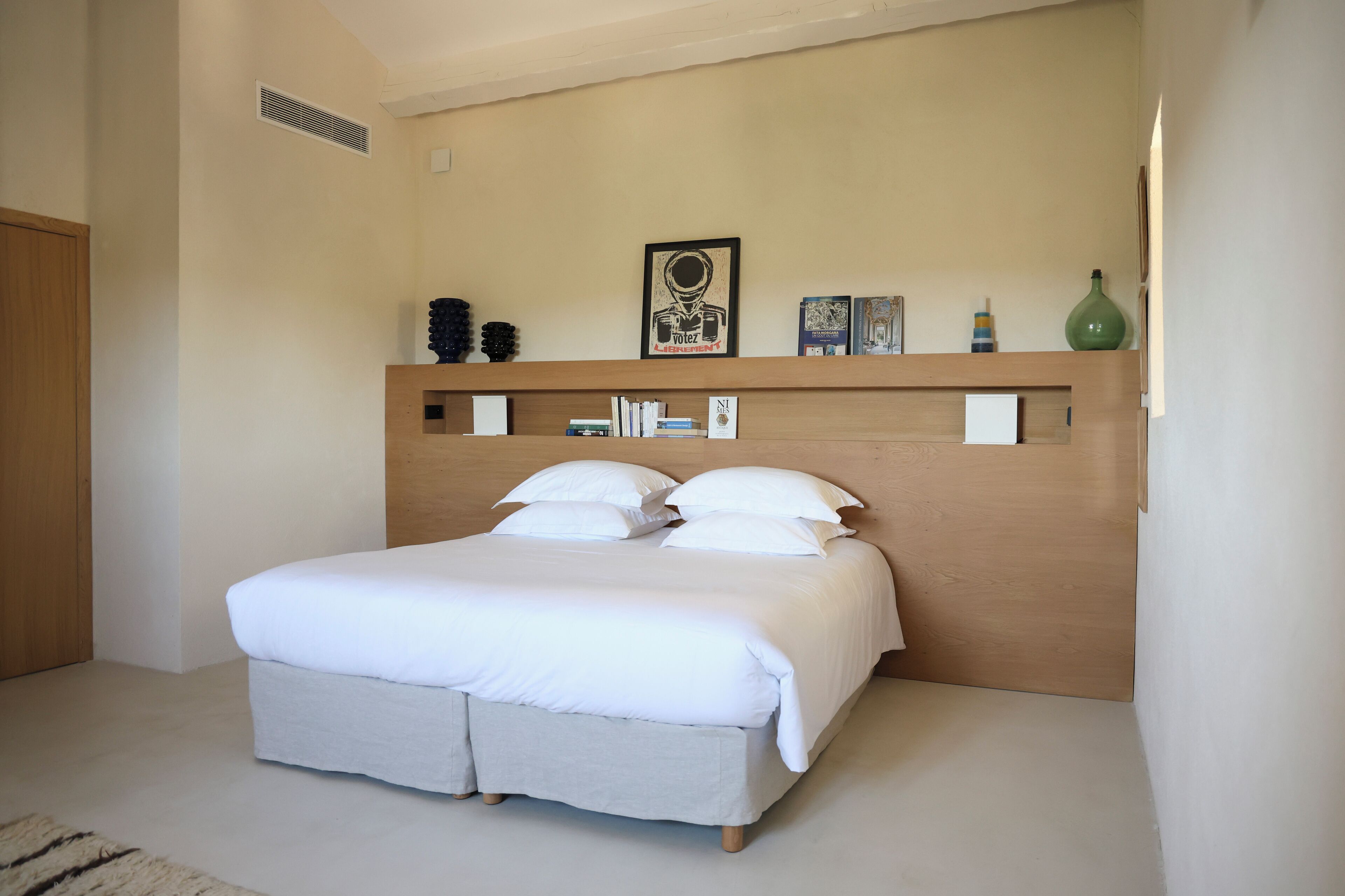 A bedroom with a large bed, a wooden headboard, a shelf with books and decorative items, and a framed picture on the wall.