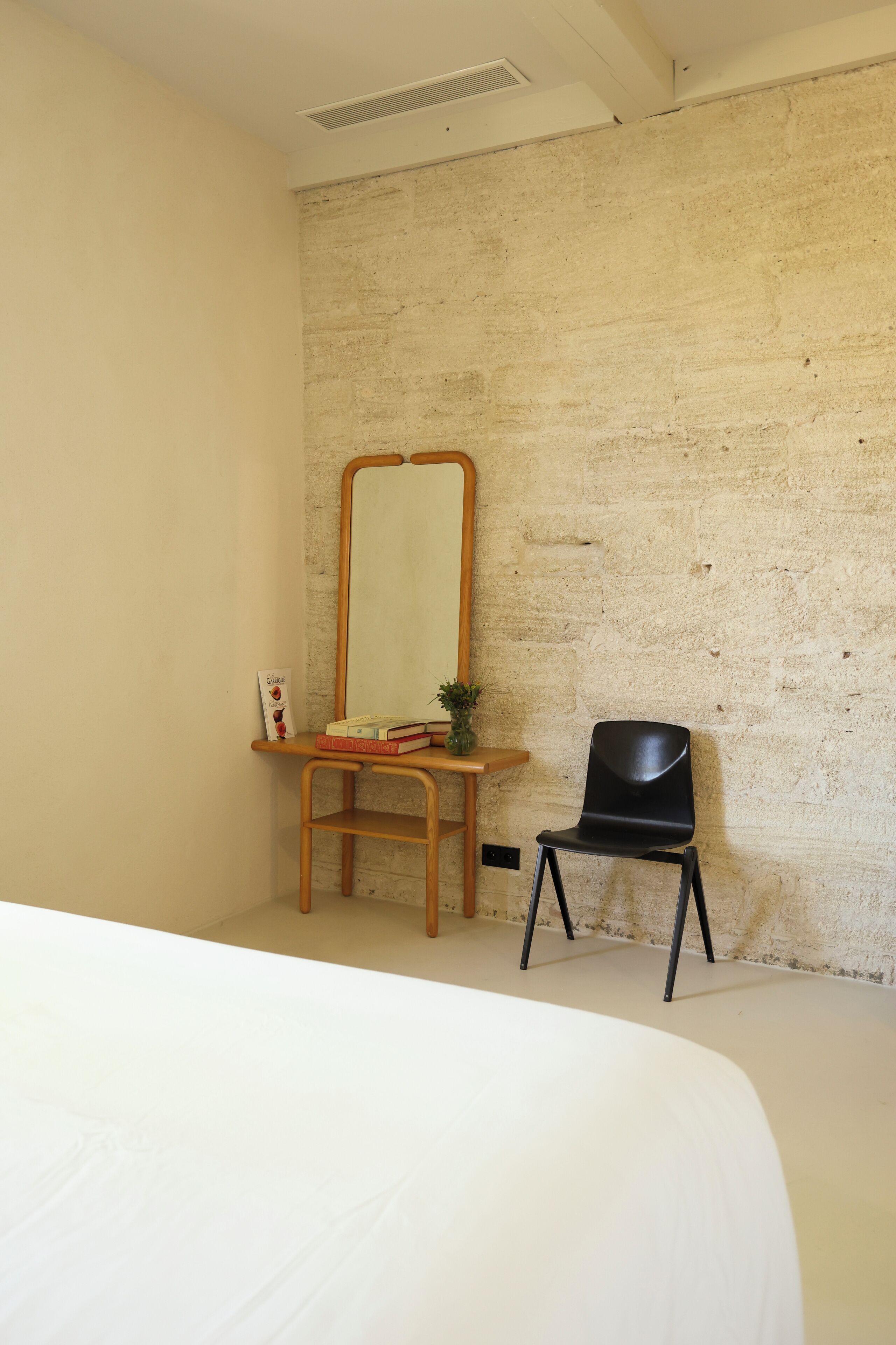 A room with a bed, a wooden desk, a black chair, a mirror, and a small plant.