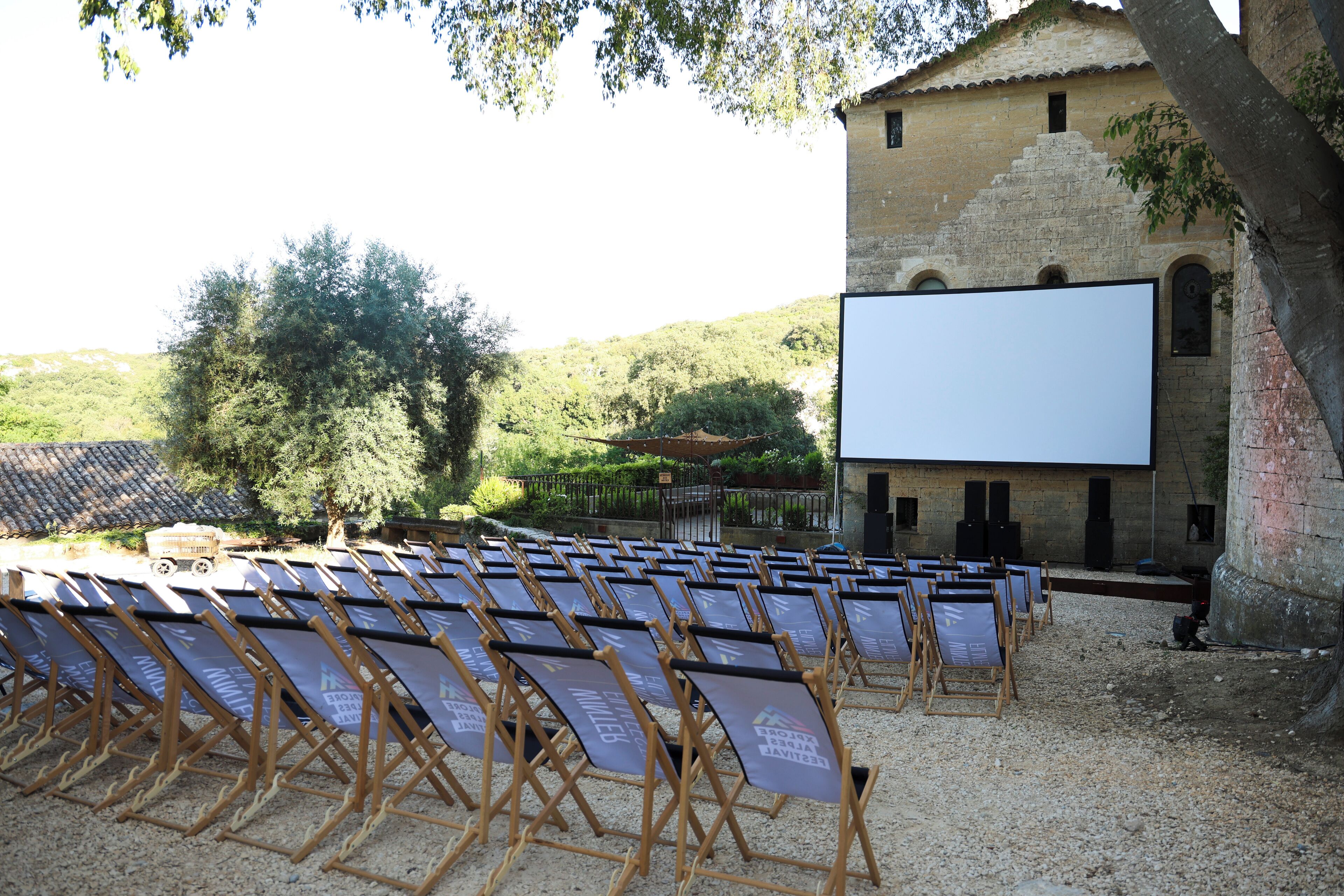 Rows of foldable chairs with a blank screen in front, set up outdoors with a building in the background.