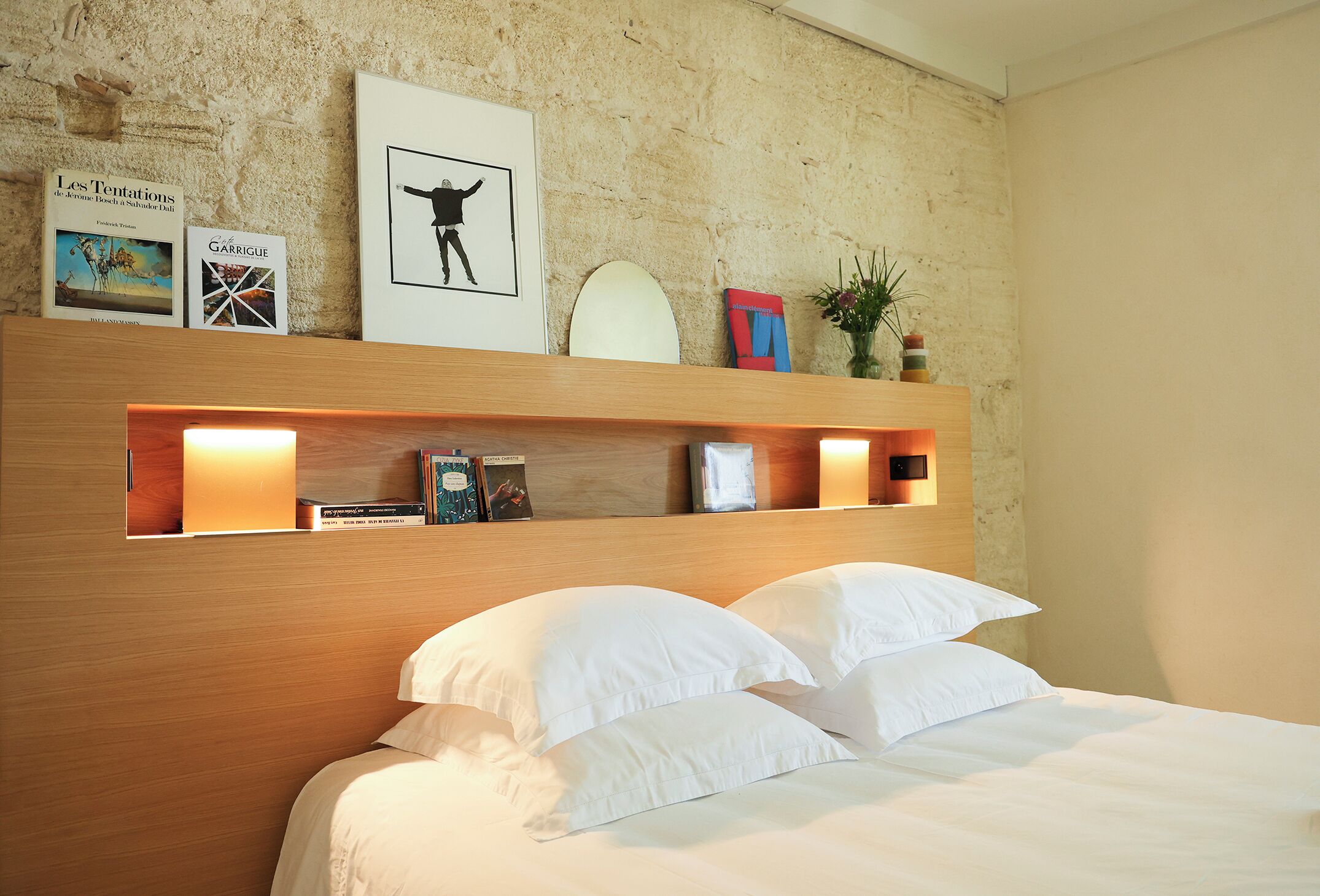 Une chambre à coucher avec une tête de lit en bois, du linge de lit blanc et une étagère contenant des livres et des objets décoratifs.