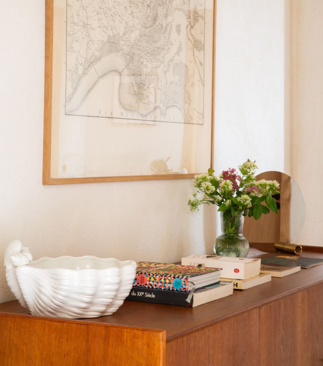 Un escritorio de madera con un cuenco blanco de cerámica, un libro, un florero con flores y un mapa enmarcado en la pared.